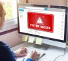 Mann sitzt vor PC-Bildschirm auf dem eine rote Warnmeldung mit "System Hacked" angezeigt wird
