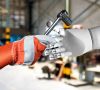 Eine behandschuhte Arbeiterhand reicht einen Ratsche einem Roboterarm - ein Symbol für die Hlfestellung, die die KI schon heute in der Instandhaltung leisten kann.